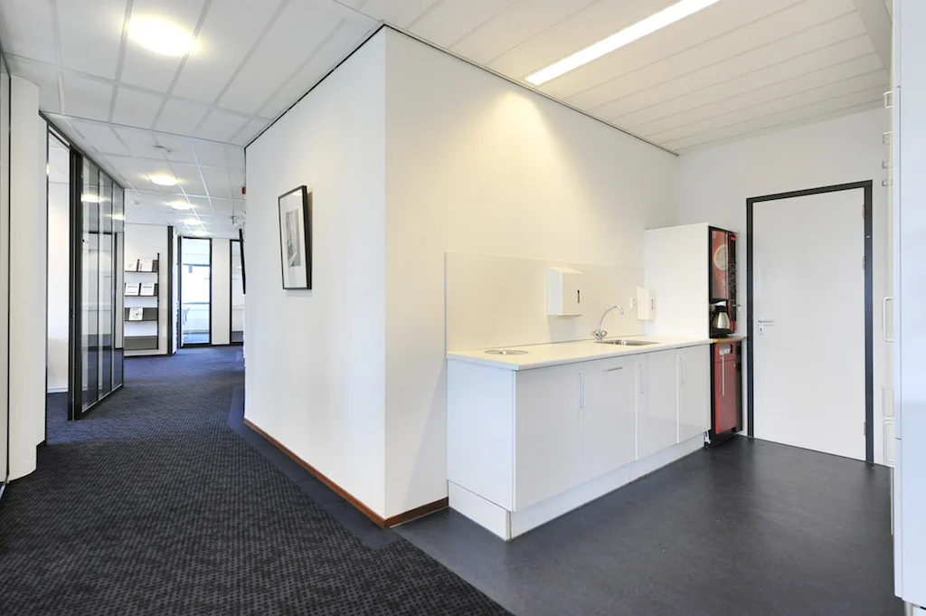 Moderne kantoorpantry met witte keukenkasten, een koffieautomaat en een gang met glazen kantoorwanden aan de Laan van S‑Gravenmade.